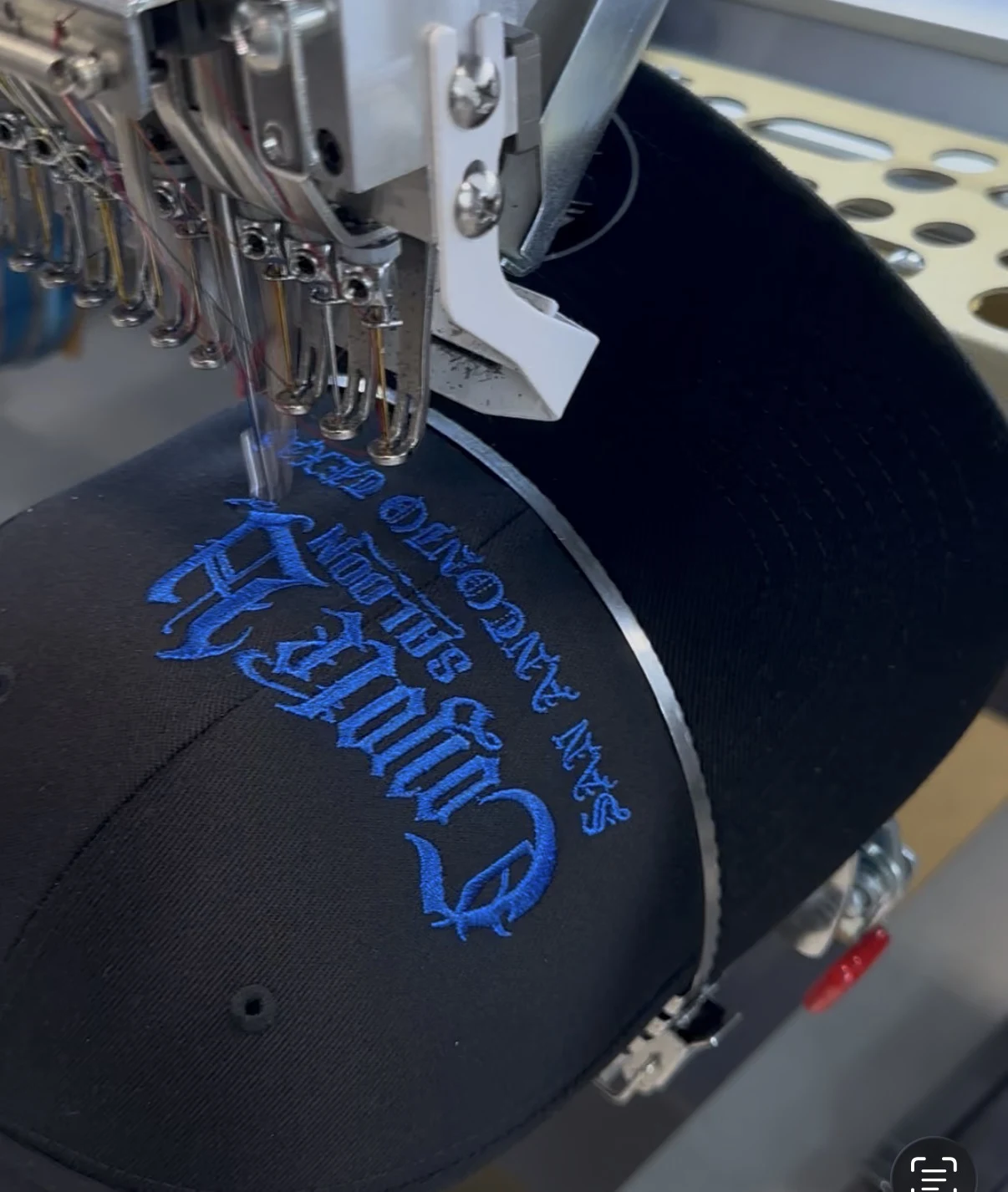 Embroidery machine stitching bright blue thread onto a custom black hat at Satellite PrintLab