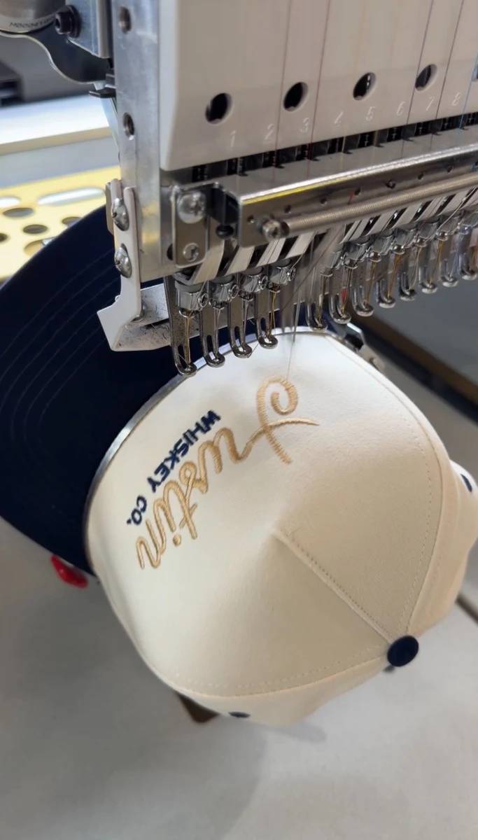 Embroidery machine stitching a cream and navy custom trucker hat at Satellite PrintLab